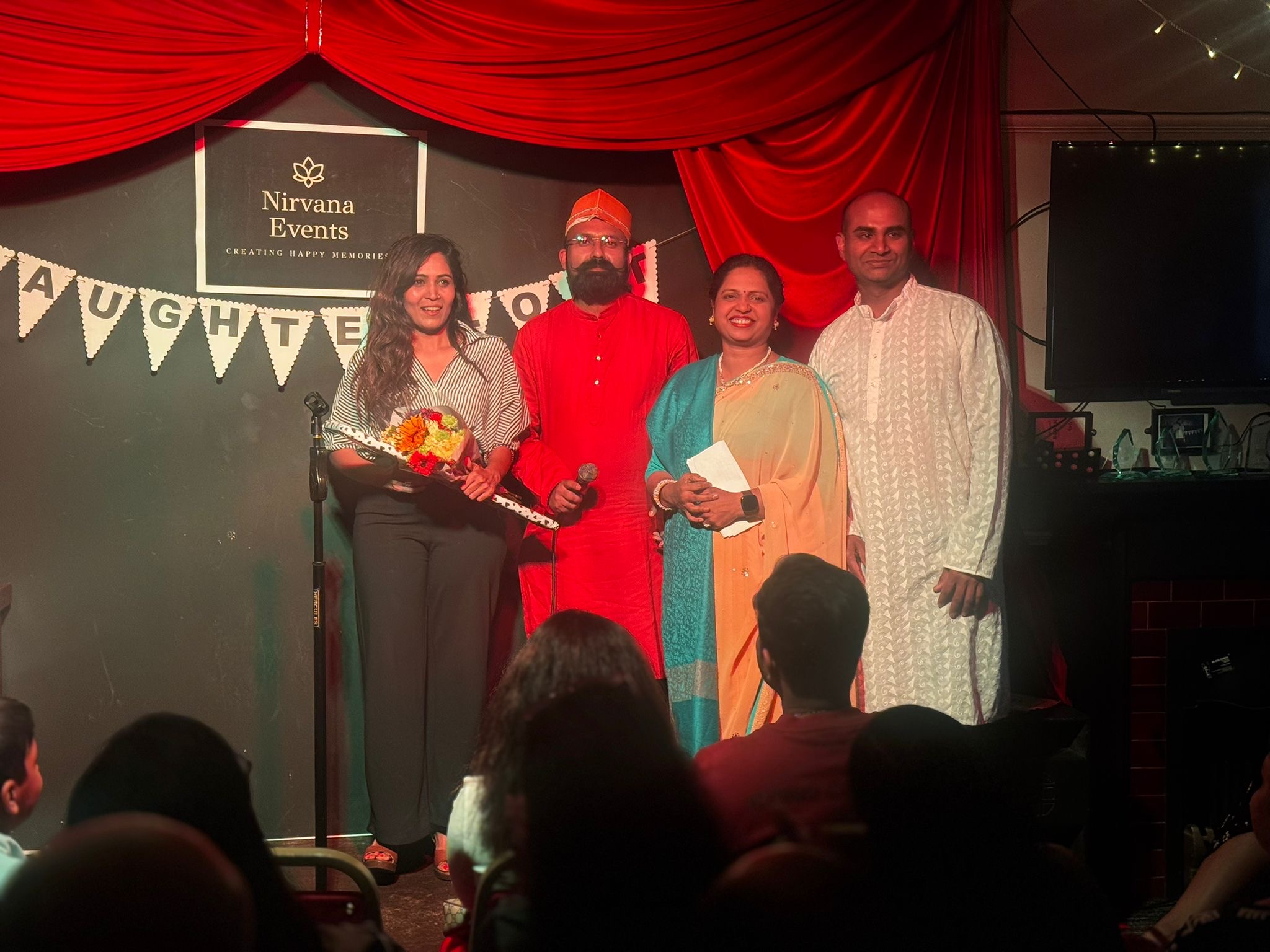 Four individuals stand on stage at the Nirvana Events venue, smiling and posing for the camera. One woman holds a bouquet of flowers, while another woman wears a colourful outfit and holds a piece of paper. The background features red drapery and a banner that reads 'Nirvana Events'.