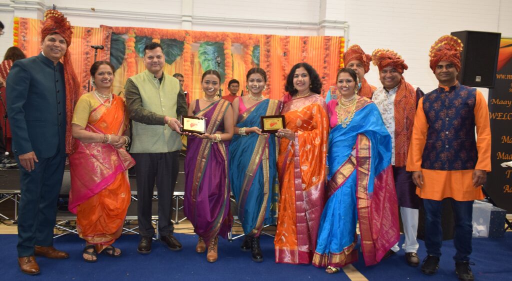 A group of people in traditional Indian attire posing for a photo, with two individuals holding a plaque in the centre. The background features colourful decorations and a stage setup.