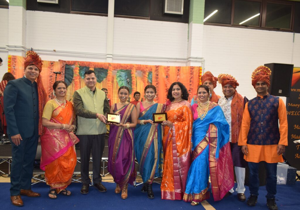 A group of eight individuals dressed in traditional Indian attire pose for a photo at a cultural event, holding awards and smiling. The backdrop features vibrant decorations, with orange and green elements.