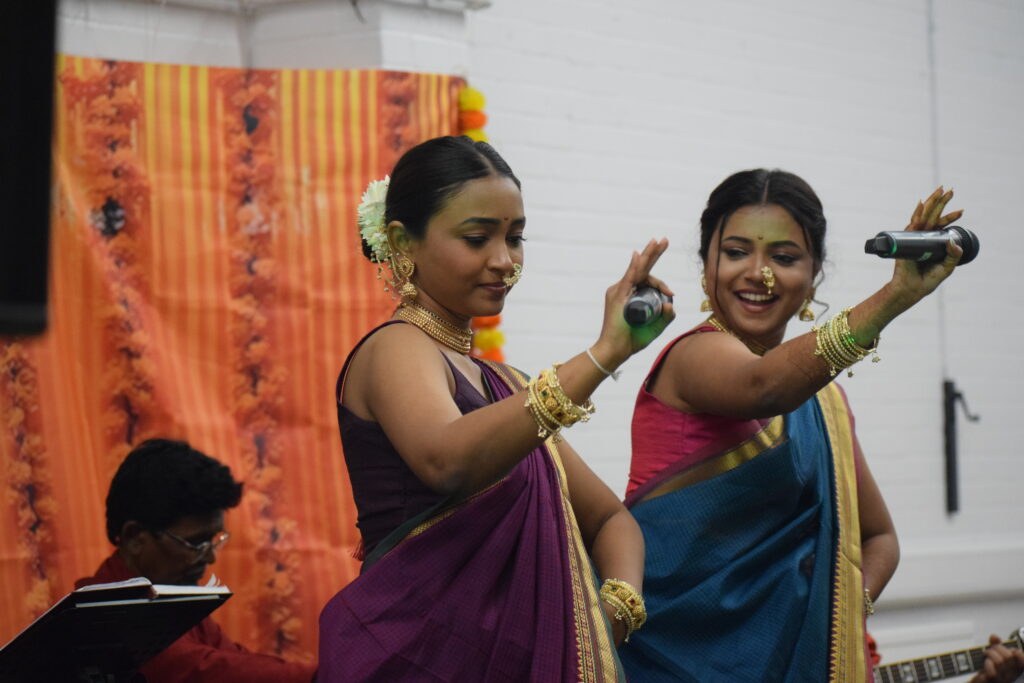 Two women in traditional attire performing a cultural dance with microphones in hand, set against a vibrant orange backdrop.
