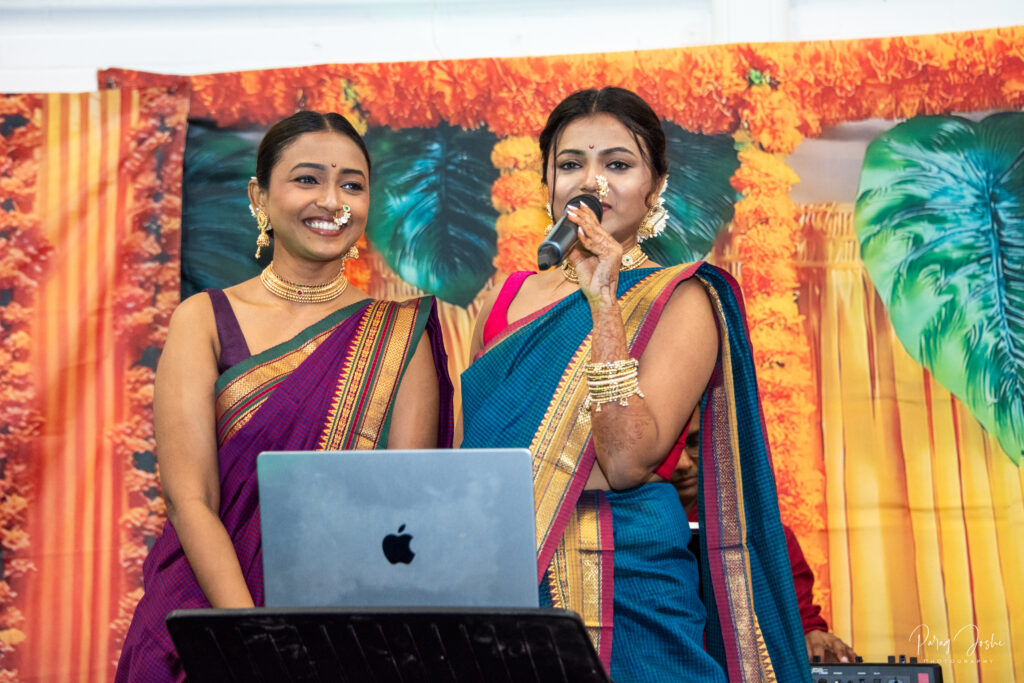 Two women in traditional Indian attire, one speaking into a microphone while the other smiles, with a decorative backdrop featuring vibrant patterns.