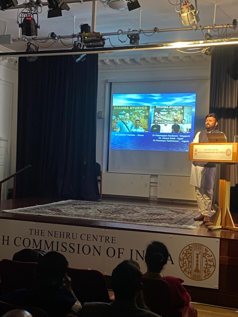 A speaker presenting at the Nehru Centre, High Commission of India, with a screen displaying information about 'Brahma Ayurved'. The audience is seated in front, and the speaker is dressed in traditional attire.