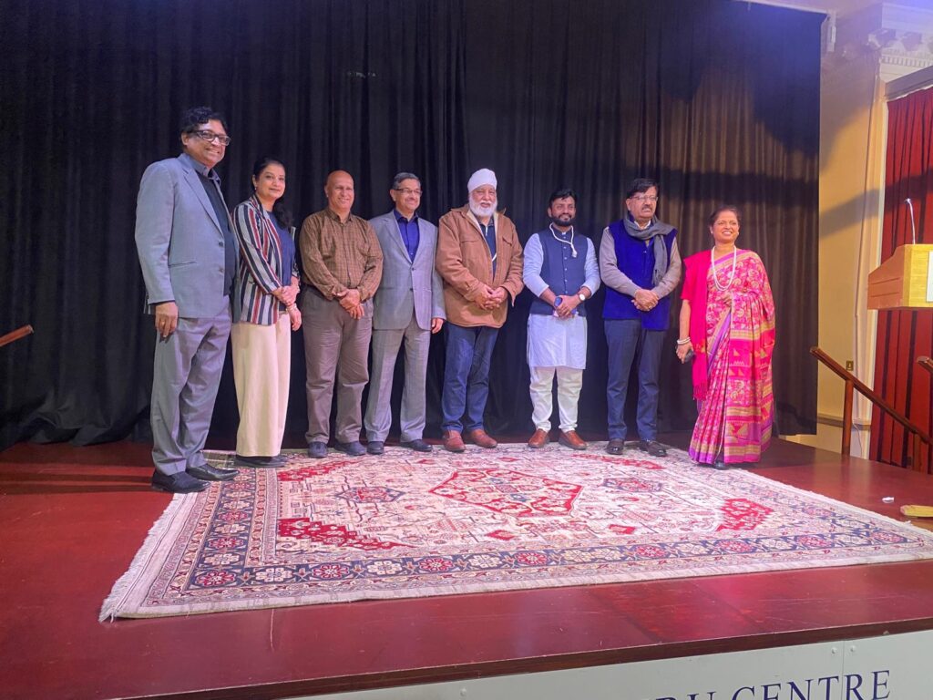 A group of eight individuals standing in a row on stage, smiling and posing for the camera. They are dressed in a mix of traditional and formal attire, with a dark curtain backdrop and a patterned carpet beneath them.